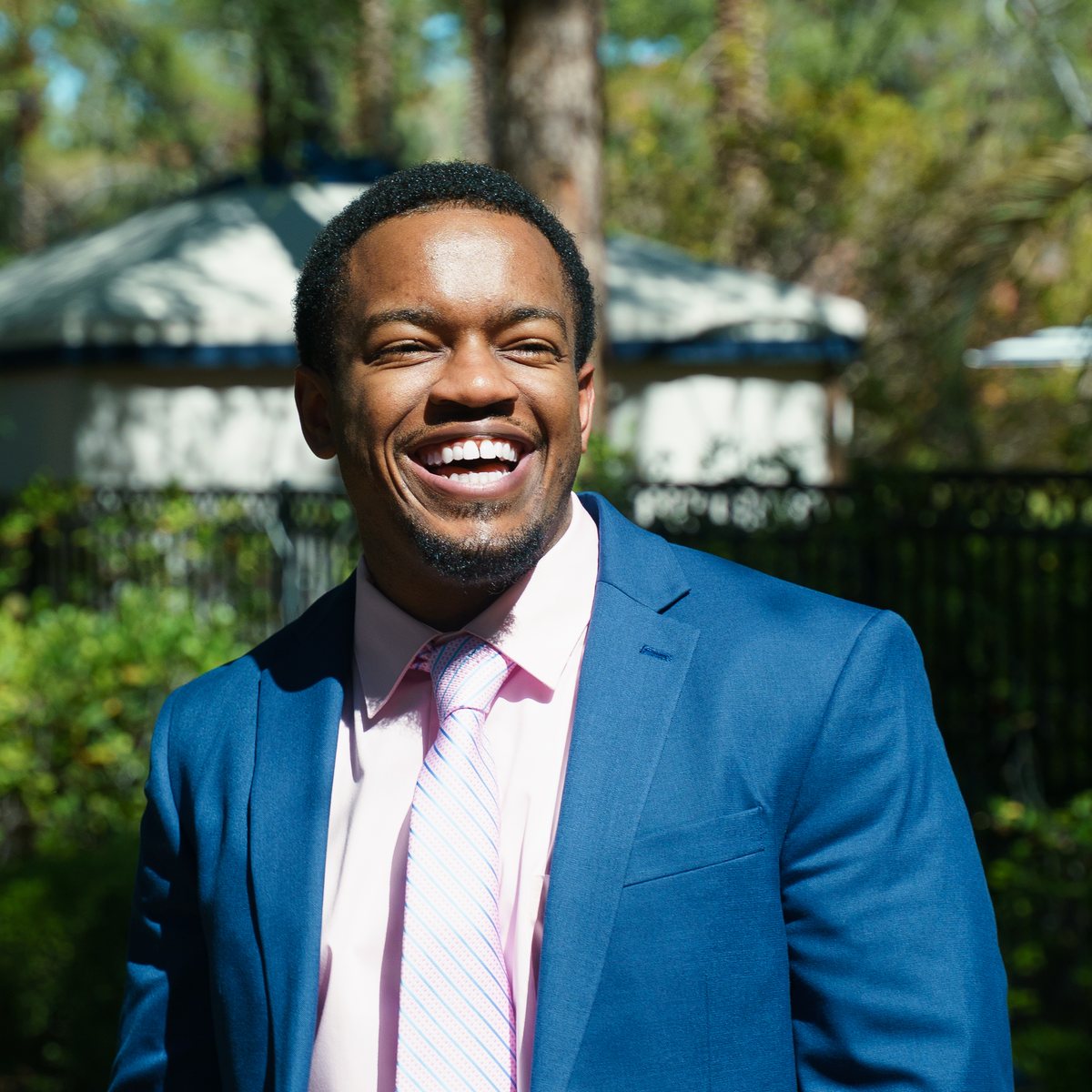 Darian Nwankwo laughing outdoors in a blue suit and pink tie